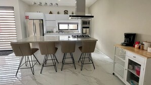 A modern kitchen with a central island, bar stools, a coffee machine, and a cabinet with kitchenware. - Casa Santacruz (Ensenada)