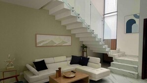 A modern living room with a white sofa, a coffee table, and a blue chair. There is a staircase with glass railings leading to an upper level. - Casa Santacruz (Ensenada)