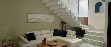 A modern living room with a white sofa, a coffee table, and a blue chair. There is a staircase with glass railings leading to an upper level.