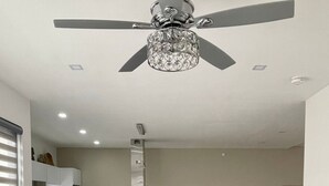 A modern dining area with a long table set for six, a ceiling fan, and a kitchen in the background. - Casa Santacruz (Ensenada)