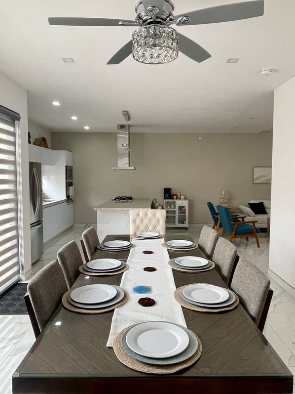 A modern dining area with a long table set for six, a ceiling fan, and a kitchen in the background. - Casa Santacruz (Ensenada)