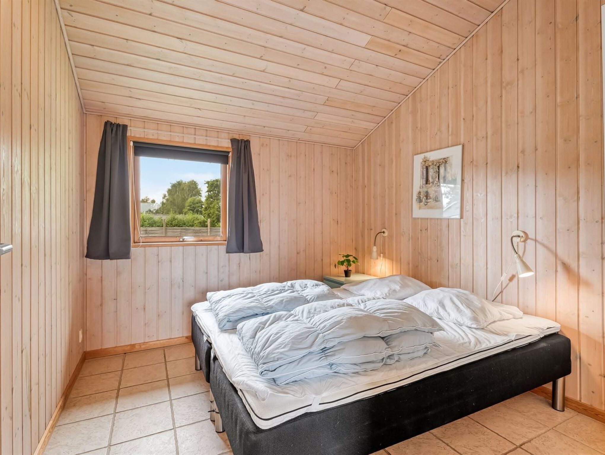 A bedroom with a wooden ceiling, a window with curtains, a bed with white bedding, a bedside table with a lamp, and a framed picture on the wall.