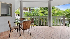A covered patio with a round table and wicker chairs, overlooking a lush garden with various tropical plants.