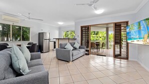A spacious living area with a grey sofa, a flat-screen TV, ceiling fans, and a view of a tropical outdoor area through a sliding door.