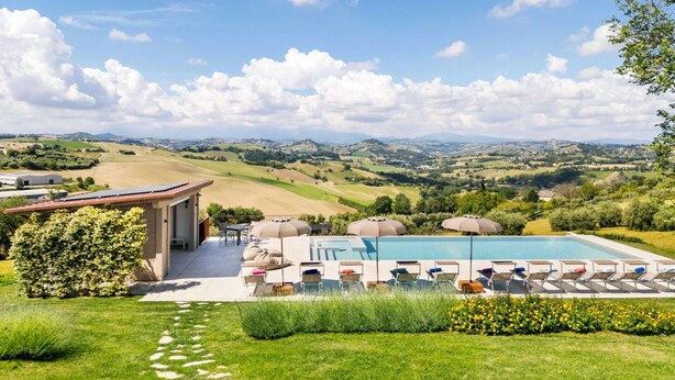 Un espace piscine avec des chaises longues et des parasols, un chemin menant à un bâtiment, et une vue panoramique sur des collines verdoyantes et une ville lointaine.