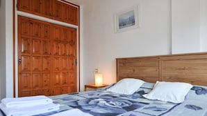 A hotel room with a wooden wardrobe, a bedside table with a lamp, a bed with a floral bedspread, and a framed picture on the wall.