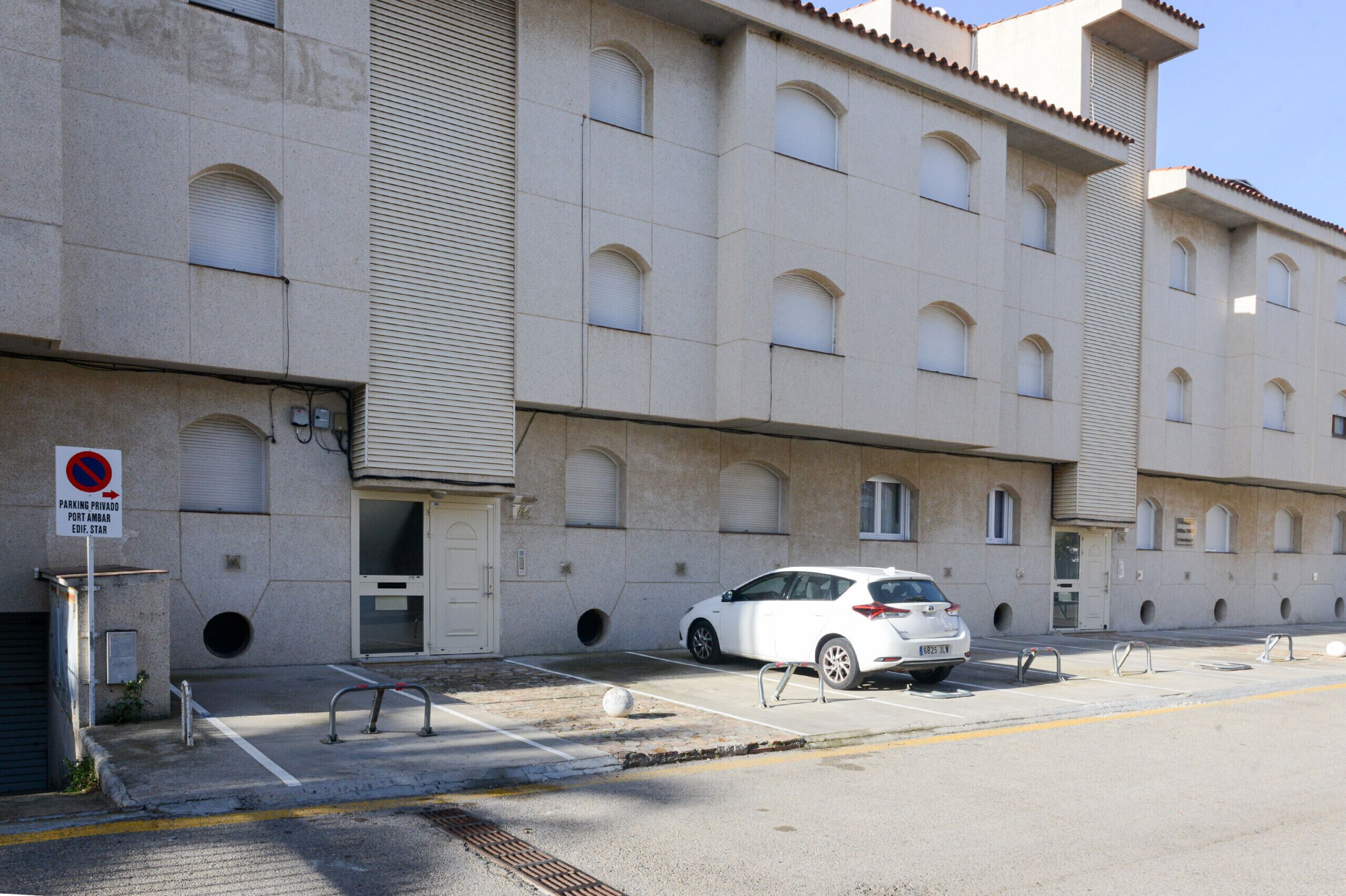 Un edificio de varios pisos con un estacionamiento, un coche blanco aparcado y un cartel de prohibido estacionar.