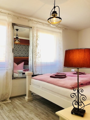 Quarto com cama, luminária de cabeceira, espelho e janela com cortinas.
