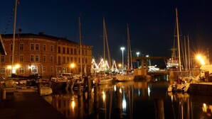 Un port de plaisance de nuit, avec des bateaux amarrés, un bâtiment doté de nombreuses fenêtres et une surface d'eau calme qui reflète les lumières.