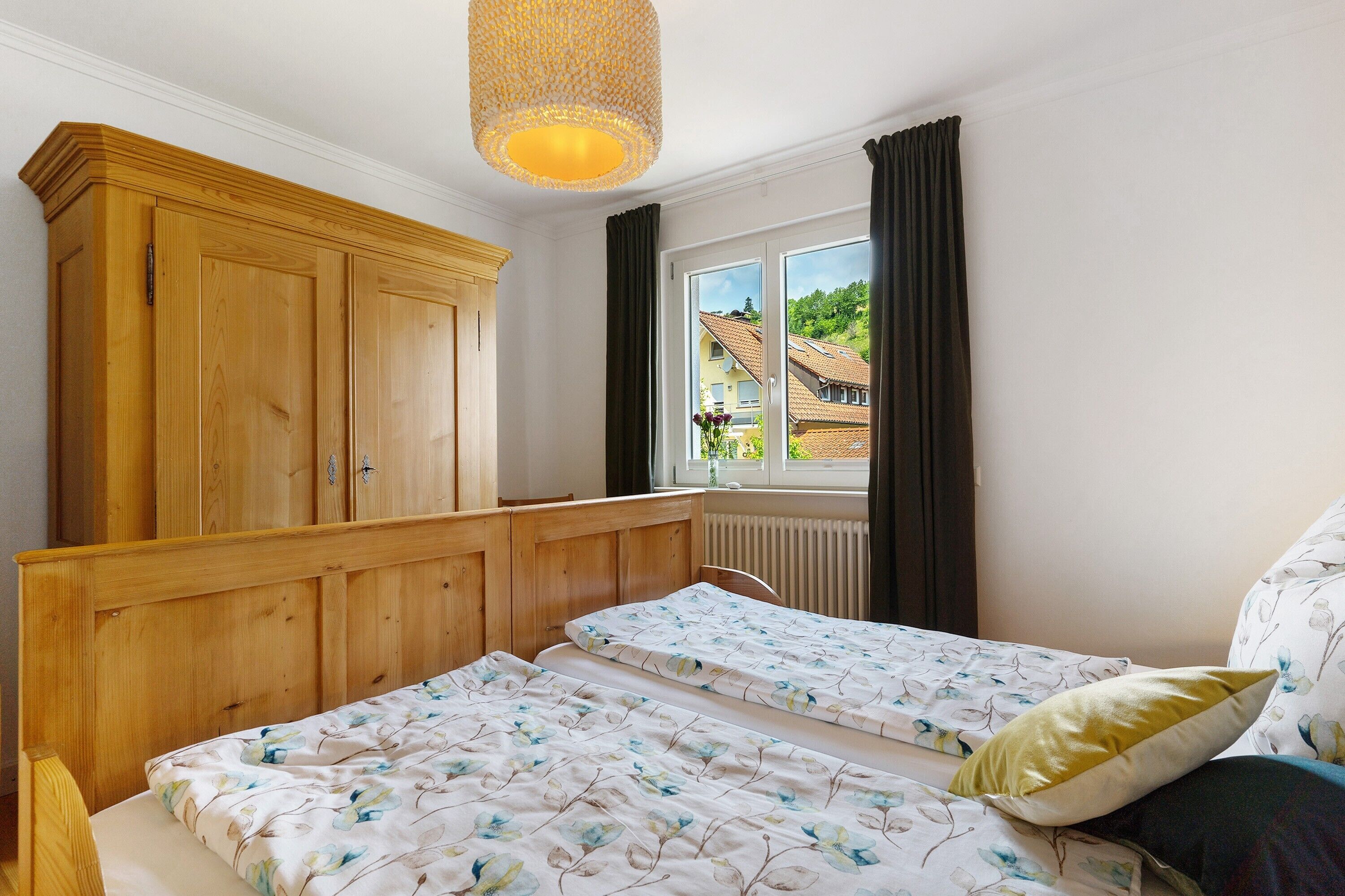 Een slaapkamer met een houten garderobe, een bed met bloemenbeddengoed, een raam met gordijnen en een hanglamp.