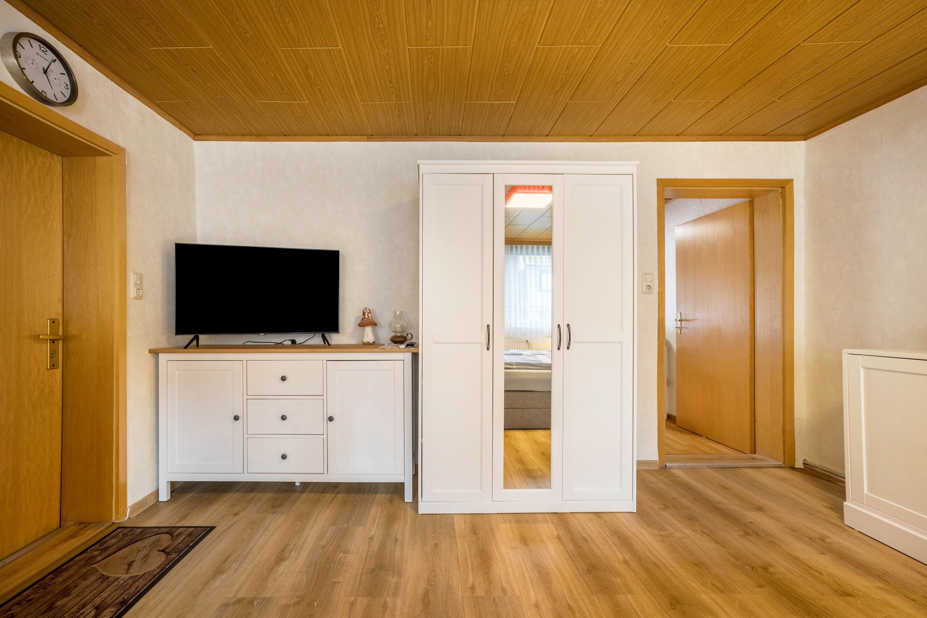 Ein Zimmer mit Holzboden, einem Fernseher auf einem Schrank, einem Kleiderschrank mit Spiegel und einer Tür zu einem anderen Raum.