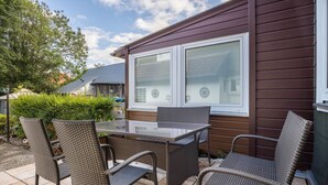 En uteplats med möbler i rotting, ett bord med glasskiva och utsikt över ett hus med brun fasad och vita fönsterramar.