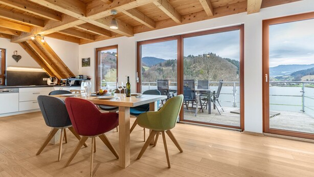 Een moderne keuken met houten balken, grote ramen en een eethoek met kleurrijke stoelen.