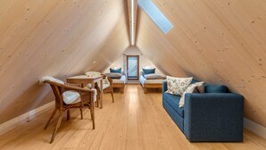Een knus zolderkamer met een schuine houten plafond, een kleine tafel met stoelen, een bank en een raam.