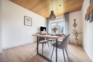 Un coin repas avec une table en bois, des chaises à revêtement gris, un luminaire suspendu, une carte encadrée, un vase avec des fleurs et une horloge au mur.