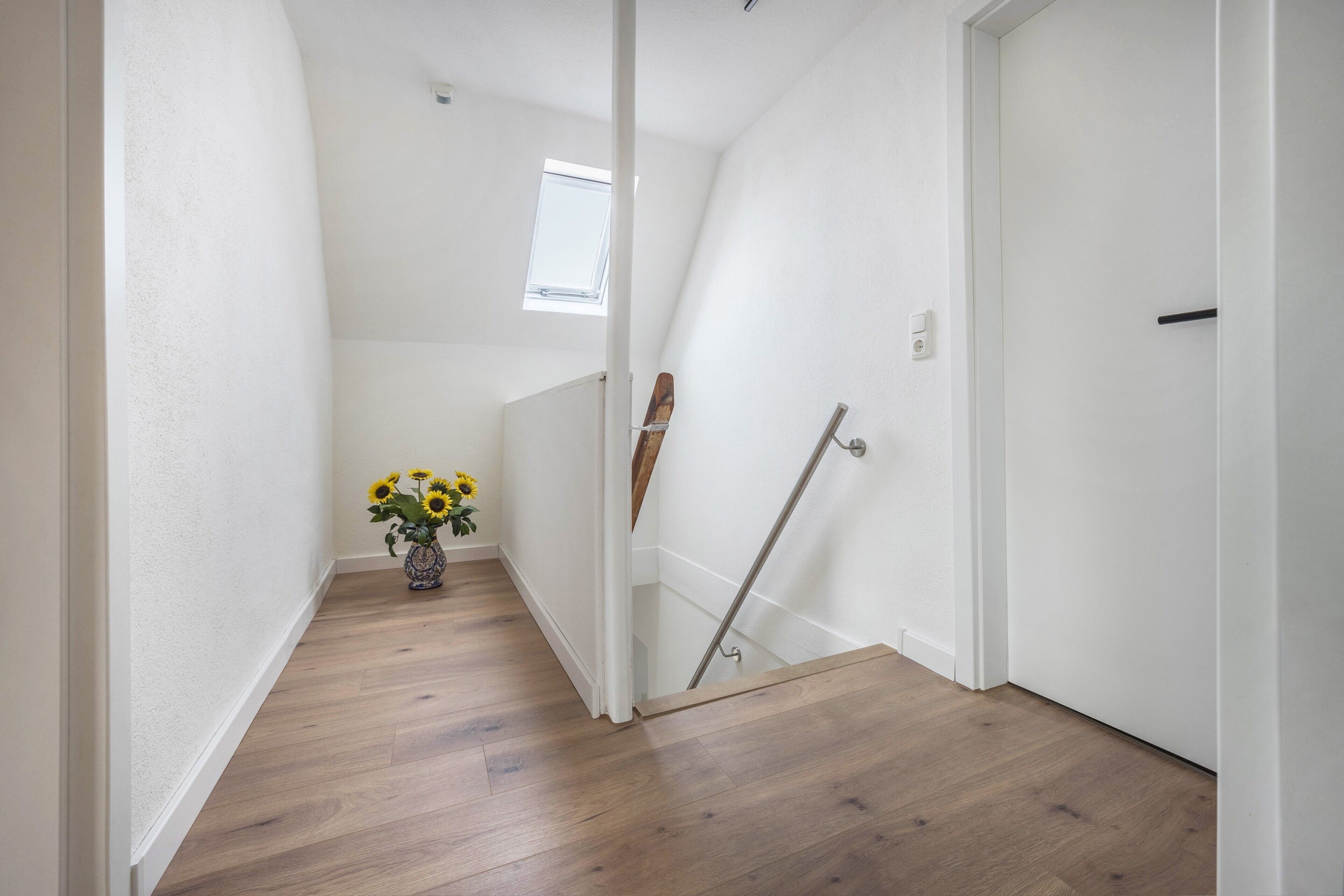 A narrow hallway with wooden flooring, white walls, a small window, and a door.