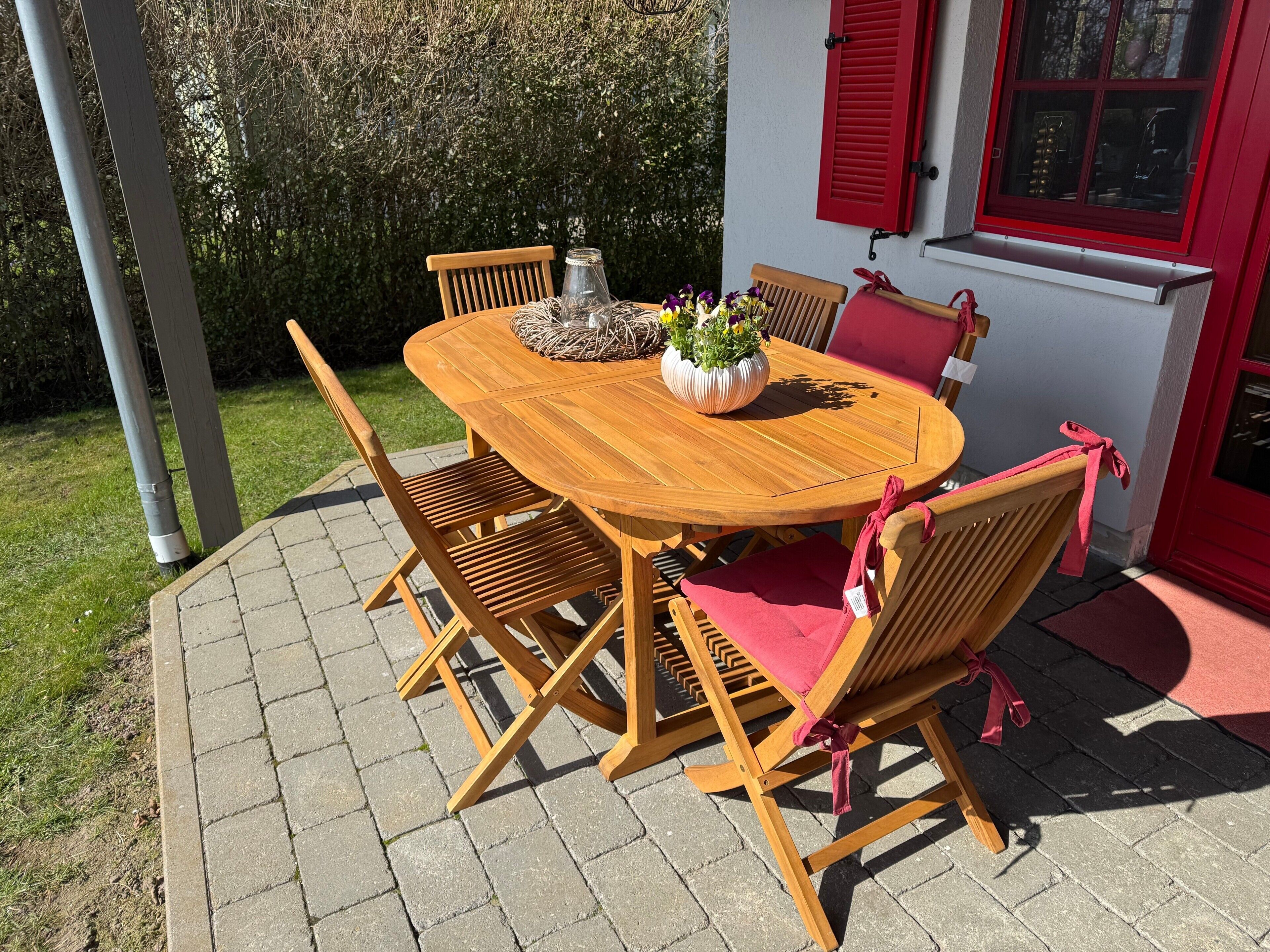 A wooden outdoor table with chairs, a potted plant, and a red window shutter.