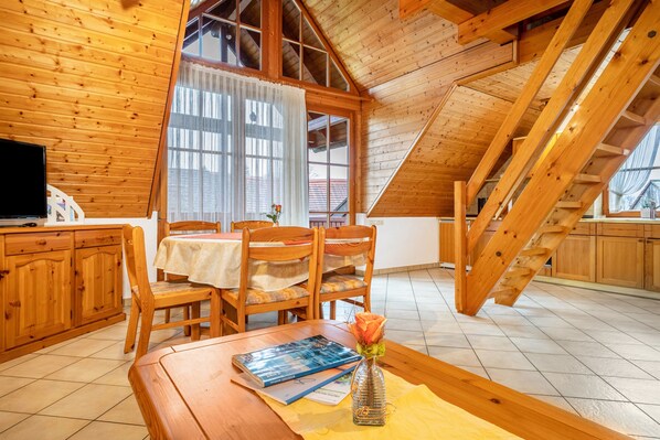 A wooden interior with a dining table, chairs, a TV, and a staircase.