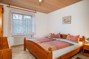 A bedroom with a wooden bed, a radiator, a window with curtains, a bedside table with a lamp, and a framed picture on the wall.