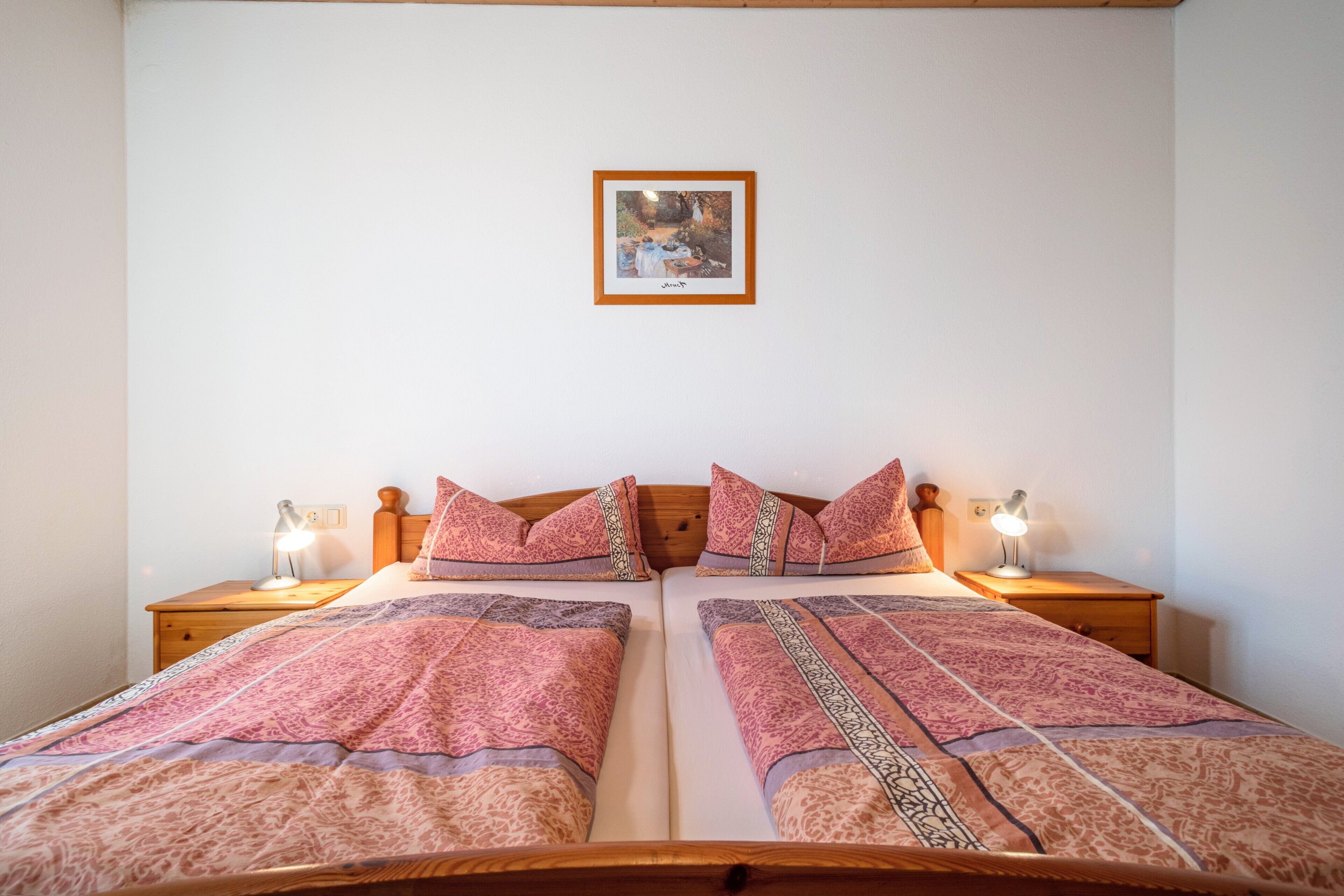 Cama doble con ropa de cama estampada, dos lámparas de noche, un cuadro en la pared y un cabecero de madera.