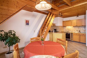 A dining area with a table, chairs, a vase with flowers, a potted plant, wooden ceiling beams, and a kitchen in the background. - Apartment 'Ferienwohnung 5, 59qm' with Shared Garden, Balcony and Wi-Fi (Kressbronn am Bodensee)