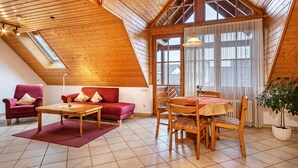 Ein Zimmer mit hohen Decken und einer geneigten Dachkonstruktion, einem Esstisch mit Stühlen, einer Couch, einem roten Sessel und einem Blumentopf.