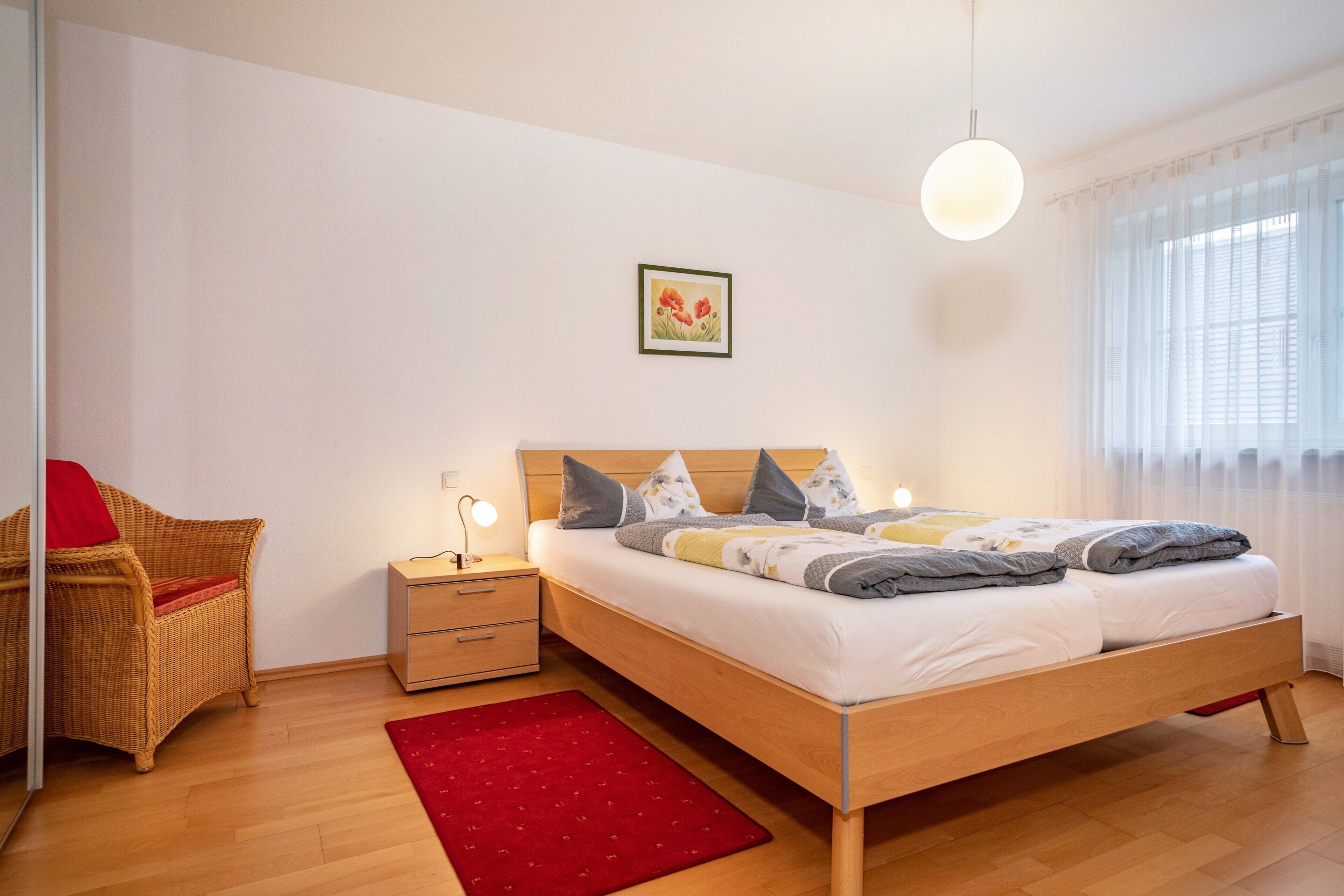 Ein Schlafzimmer mit einem hölzernen Bett, einem Nachttisch, einem roten Teppich, einem Korbstuhl und einem Fenster mit transparenten Vorhängen.
