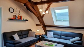 A loft-style living room with exposed wooden beams, a wooden coffee table, two sofas, a wall clock, and a skylight.