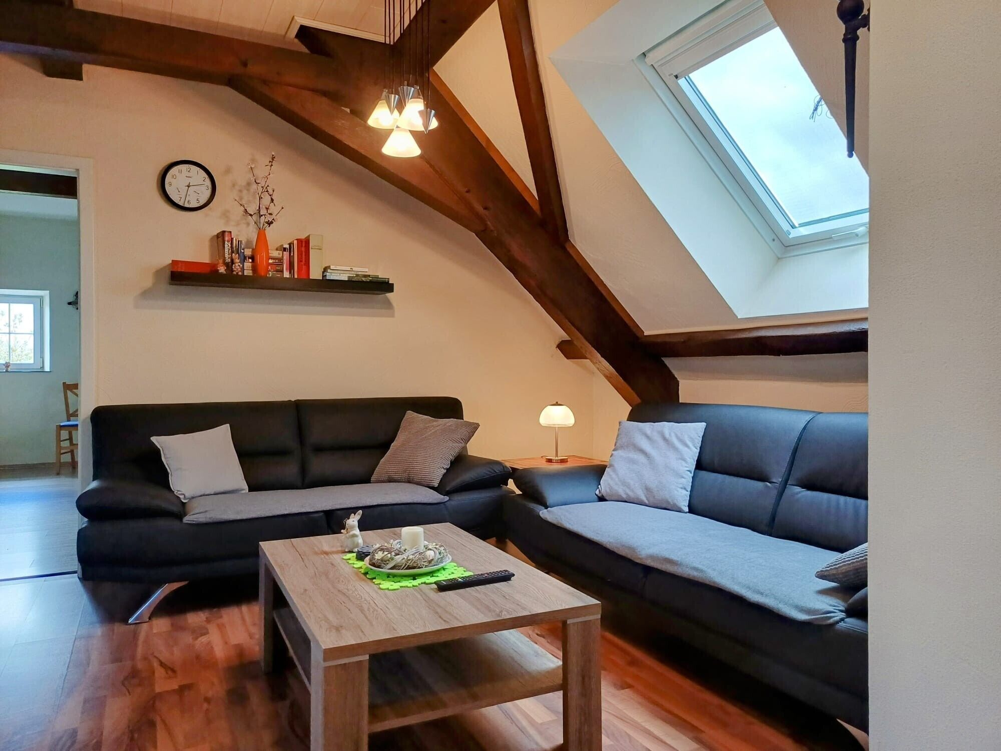 A loft-style living room with exposed wooden beams, a black sofa, a wooden coffee table, and a skylight.