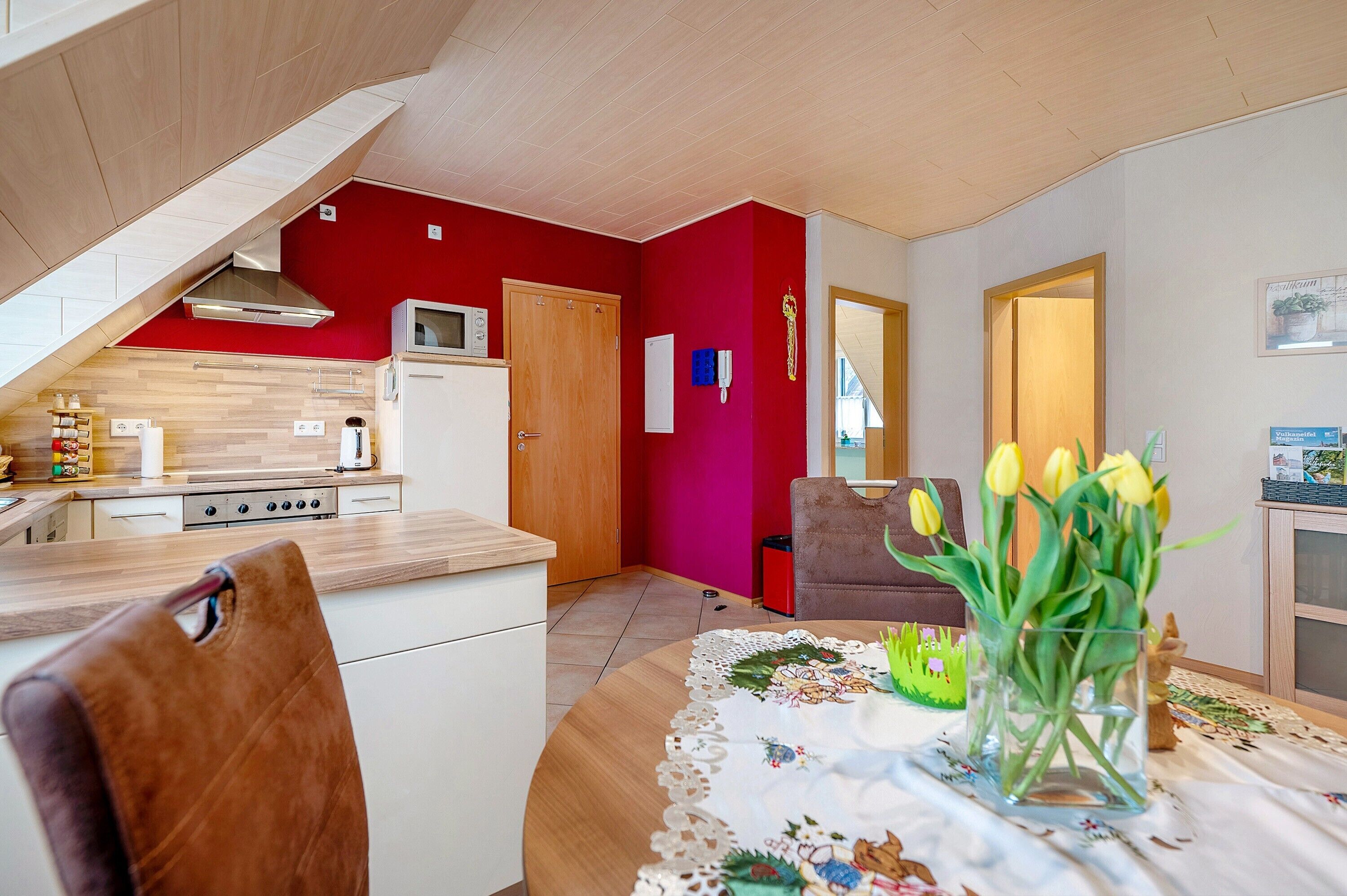 Una cocina moderna con una pared de acento roja, un comedor con una mesa dispuesta para dos personas, y vistas al salón.