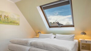 Un dormitorio con tragaluz, una cama con ropa de cama blanca, una mesita de noche con lámpara y un cuadro en la pared.