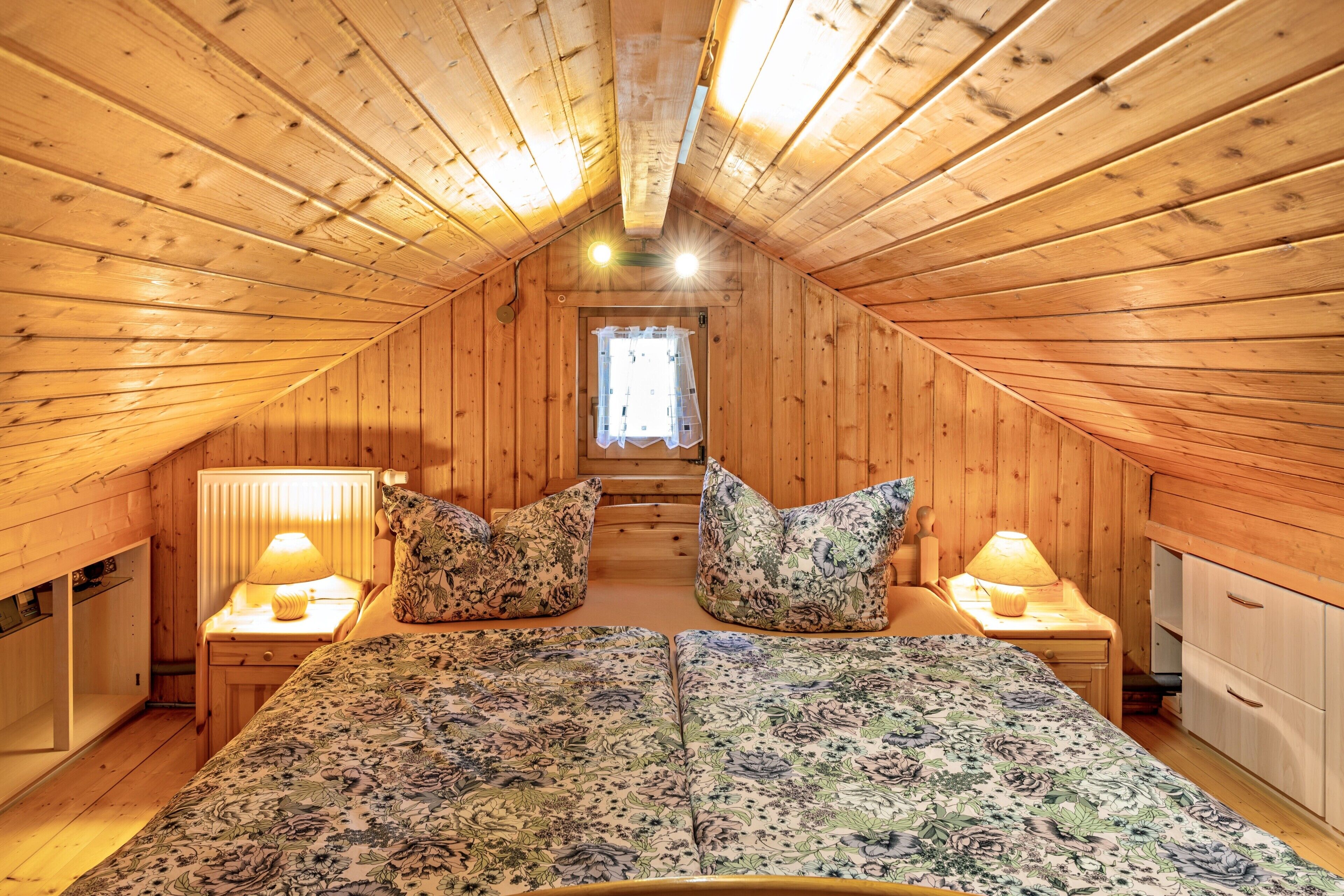 A wooden attic bedroom with a bed, two nightstands, a window, and a wooden wardrobe.