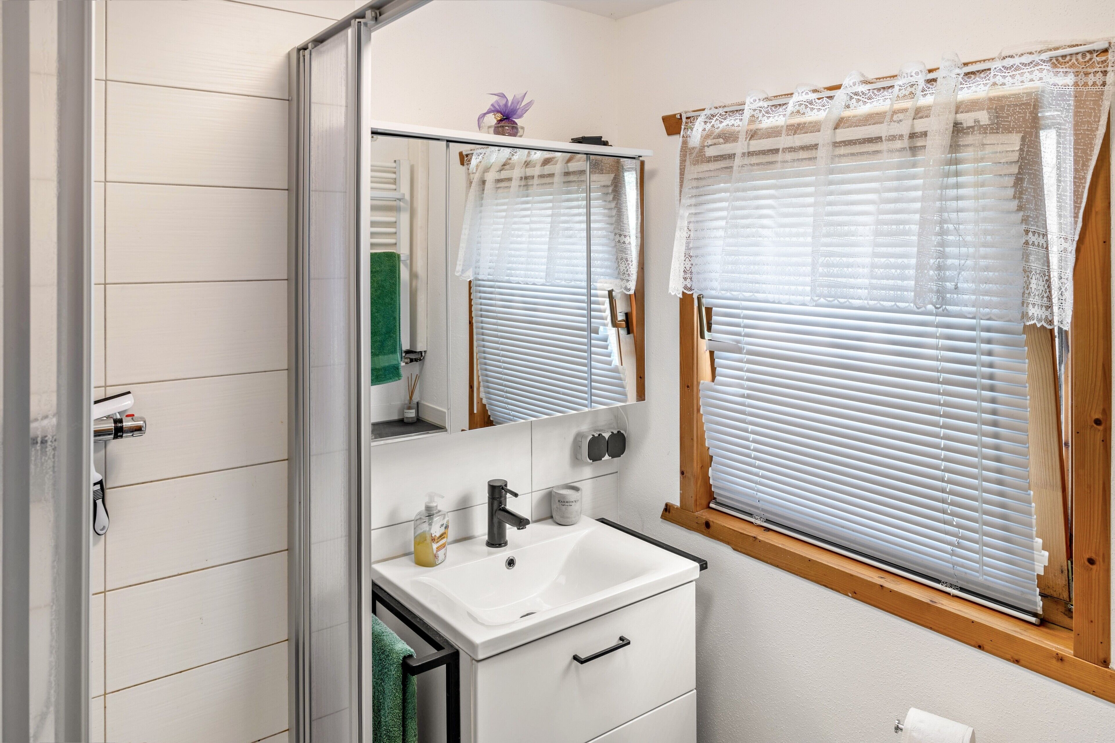 Une salle de bain avec un lavabo blanc, un miroir et une fenêtre avec des stores.