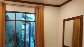 A bedroom with a bed, a ceiling fan, a window with curtains, a mirror, and a small wooden cabinet. - Oi Beach (Maamigili)