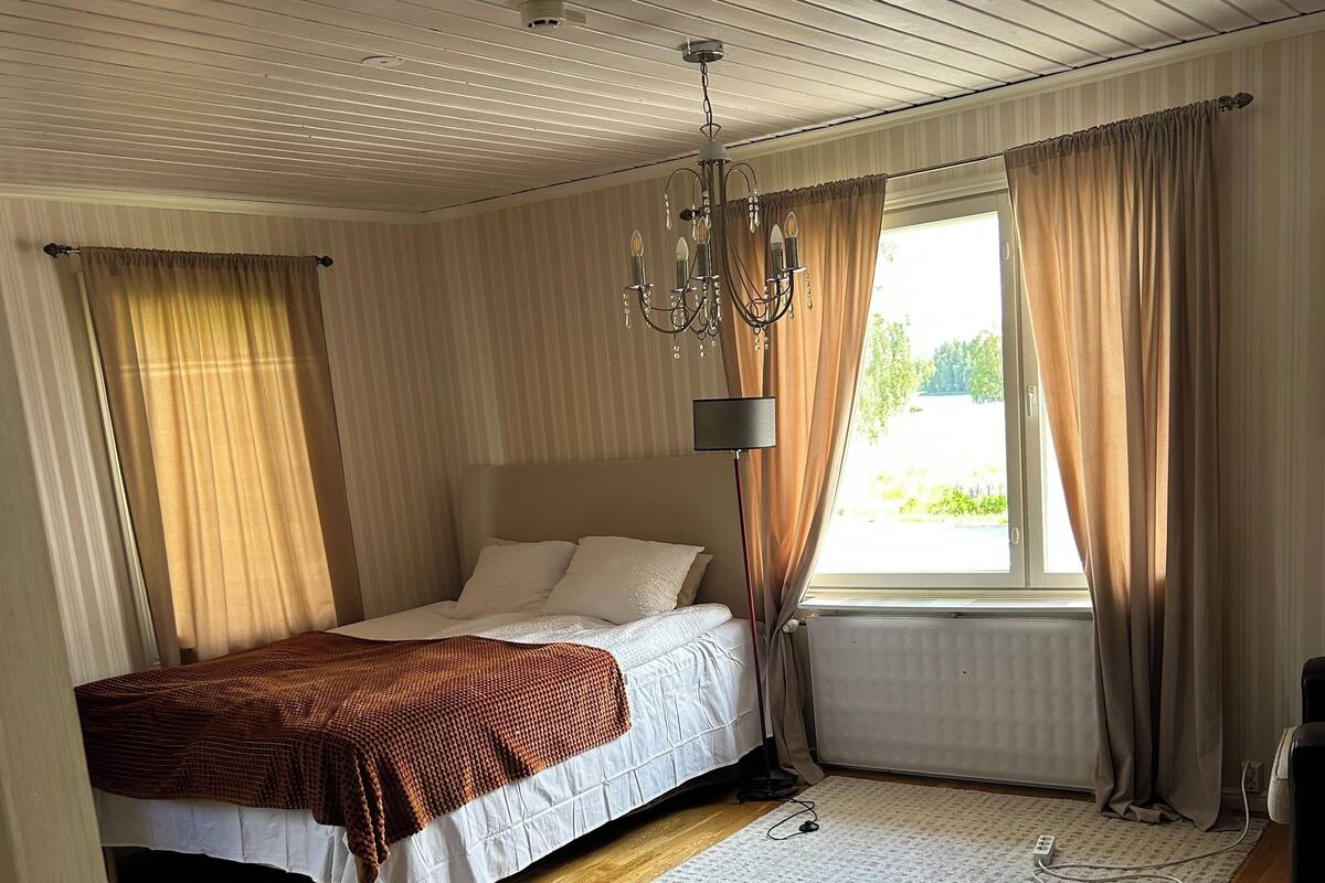 A bedroom with a bed, a standing lamp, a window with curtains, and a wooden floor.