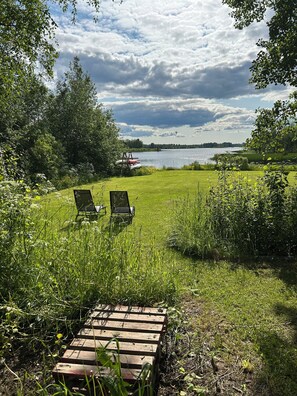 Une zone herbeuse avec deux chaises de jardin disposées face à un lac, le tout entouré d’arbres et de végétation.