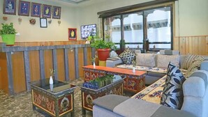A traditional room with a sofa set, a coffee table, and a TV. There are framed pictures on the wall and a window with curtains. - Pema Yangsel Hotel (Paro)