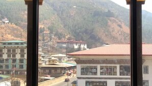 A view through a window with a mountain backdrop, featuring a road, buildings, and a temple. - Pema Yangsel Hotel (Paro)