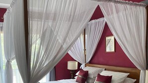 Una cama con dosel, ropa de cama blanca y almohadas rojas, un cabecero de madera y una mesita de noche con lámpara.
