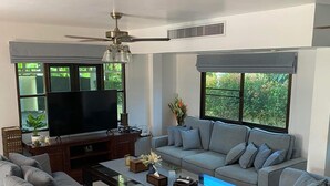 Una sala de estar moderna con un televisor de pantalla plana, sofás grises, una mesa de centro y ventanas grandes con persianas.