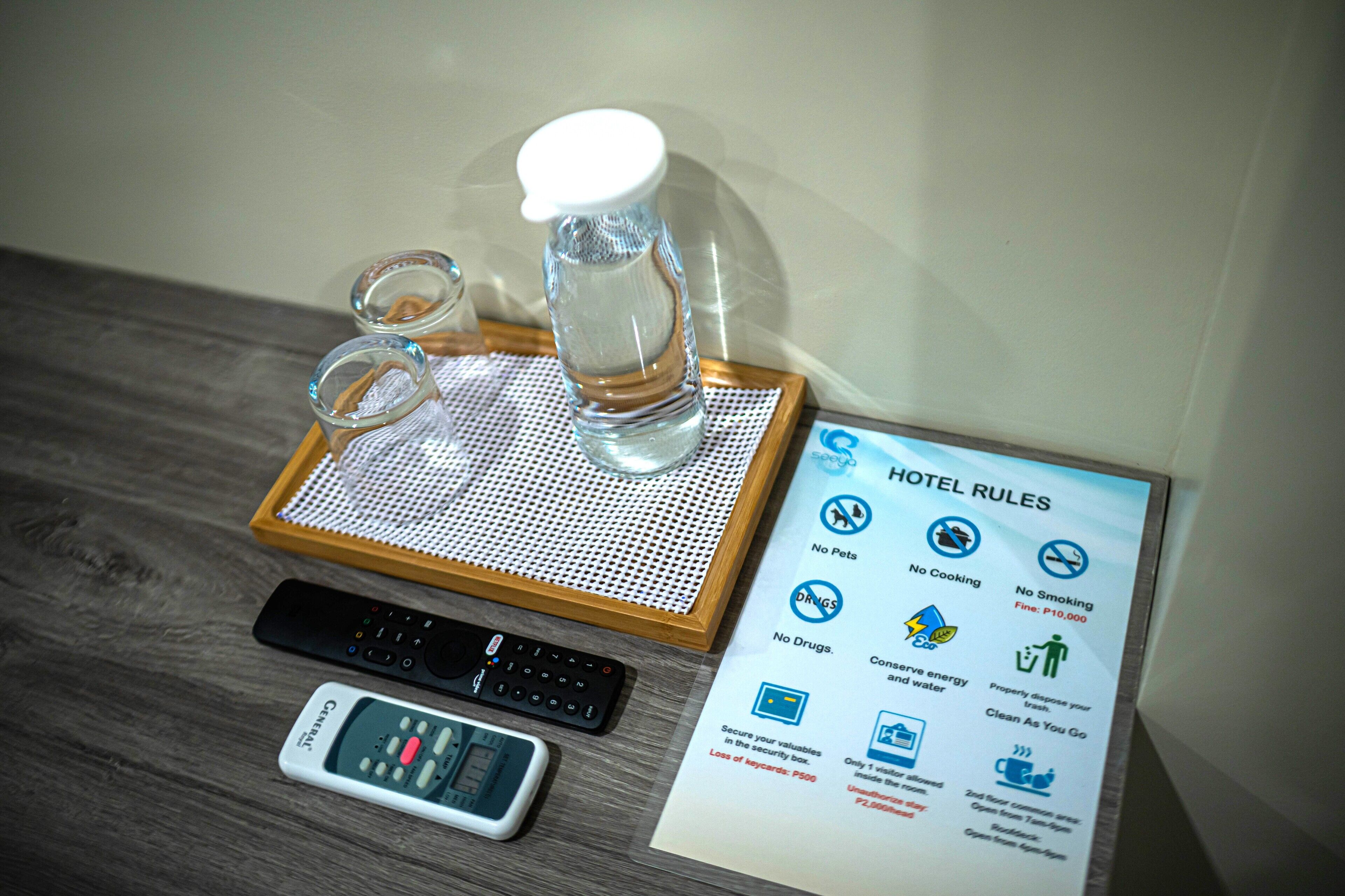 Habitación de hotel con un control remoto, un teléfono inteligente, un vaso de agua y una bandeja con un vaso y una botella. Se ve un cartel con las normas del hotel.