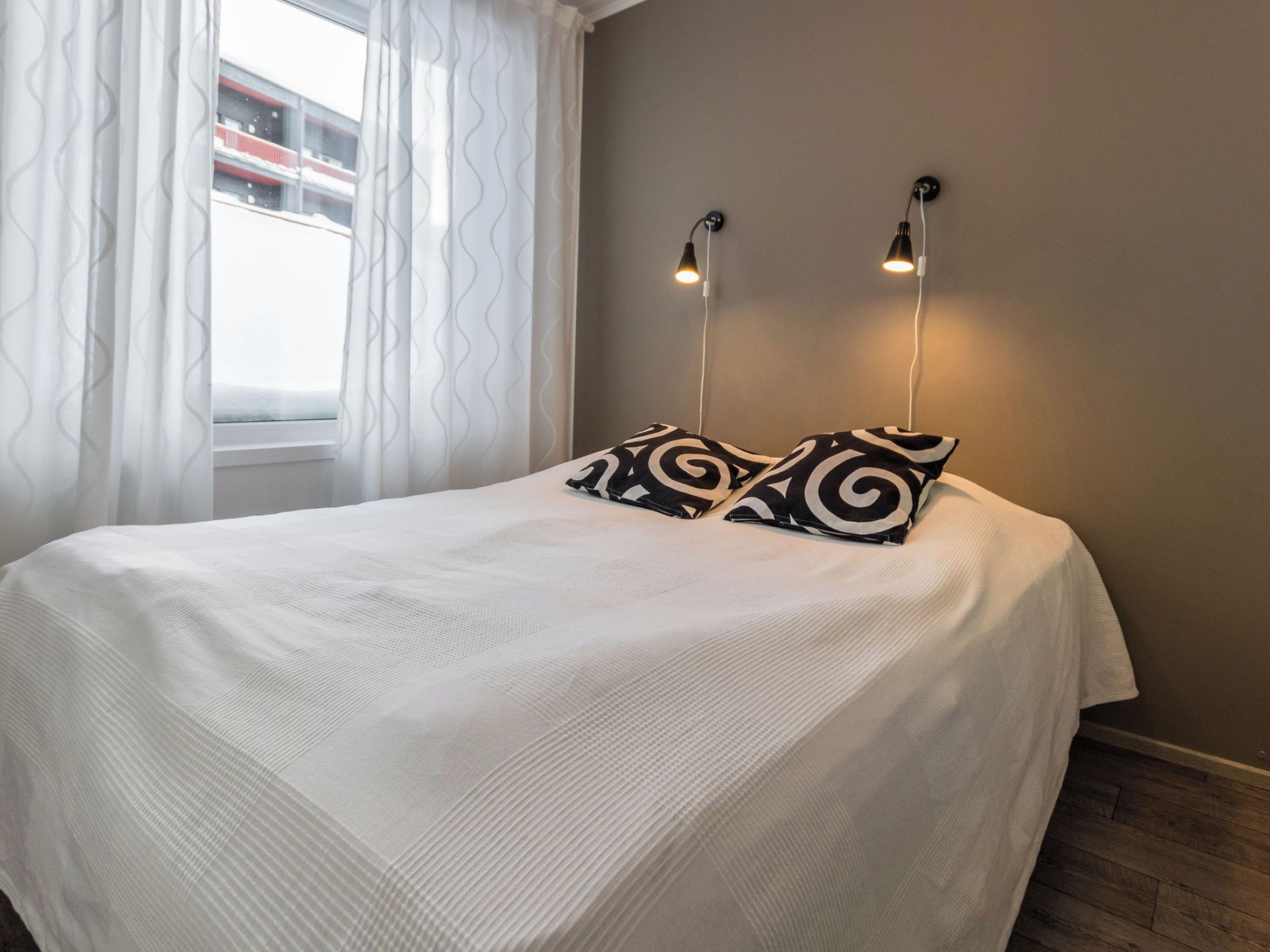 A bed with a white bedspread and two black and white patterned pillows. There are two wall-mounted lights above the bed.
