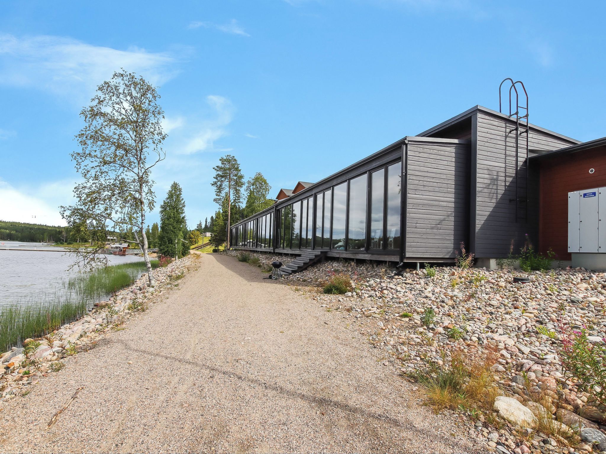 Un chemin de gravier mène à un bâtiment moderne doté de grandes fenêtres, d’une terrasse en bois et offrant une vue sur un lac.