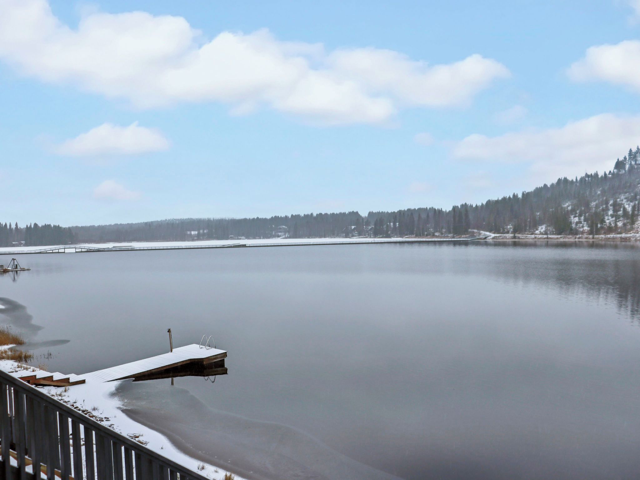 Tyyni järvi, jonka rannalla on laituri, joka ulottuu veden päälle. Järveä ympäröi lumipeitteinen maa ja metsäinen rinne.