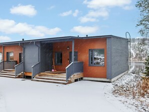 En modern byggnad med en inglasad veranda, trappsteg som leder upp till entrén och utsikt över en sjö i bakgrunden.
