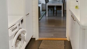Un cuarto de lavado con lavadora y secadora de carga frontal, piso de madera y, al fondo, un comedor con mesa y sillas.