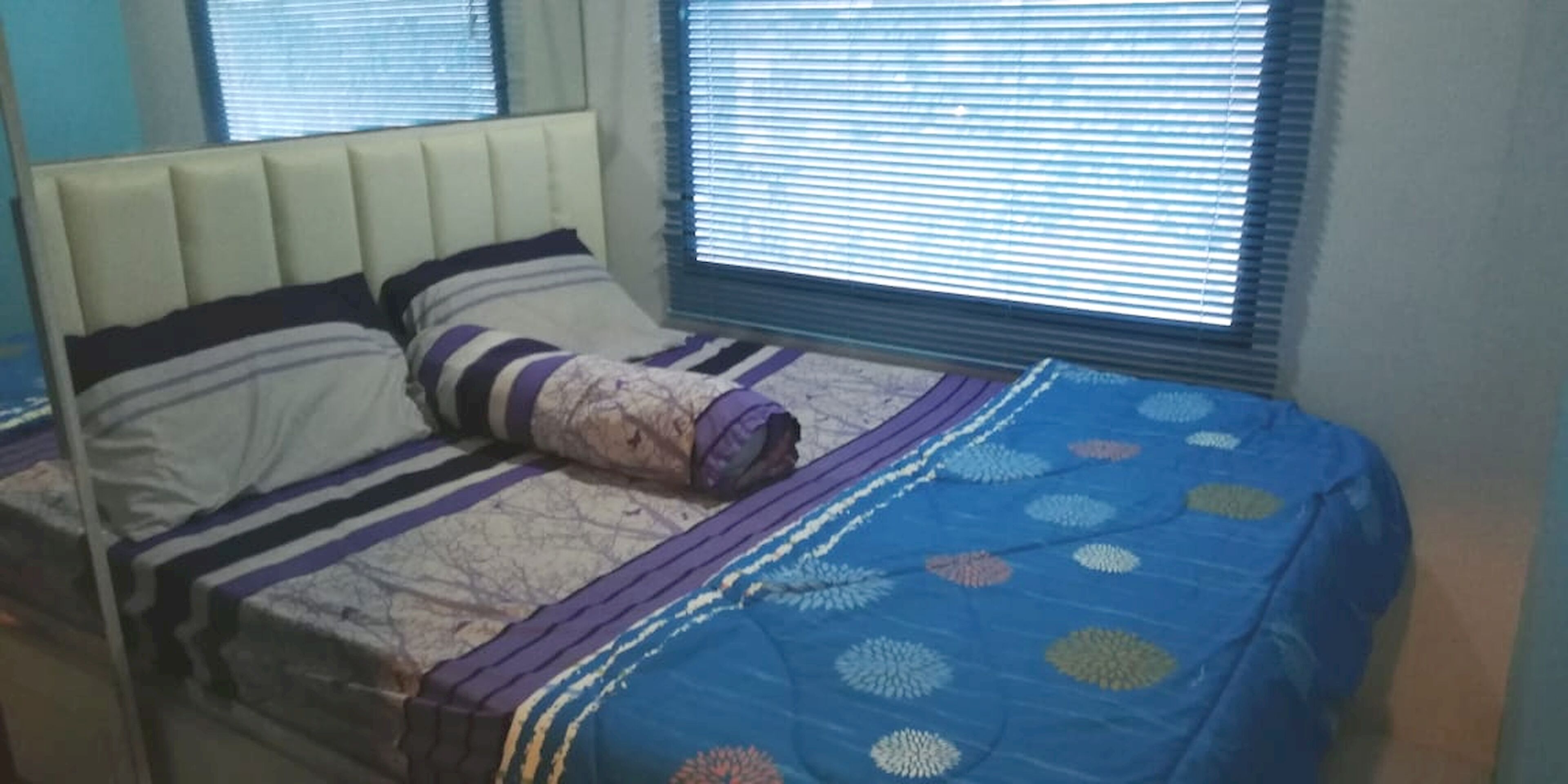 A bedroom with a bed, a headboard, and two windows with blinds.