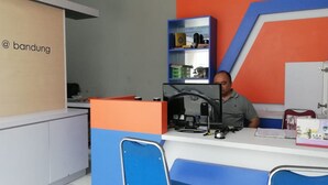 A reception desk with a man seated behind it, a blue and orange color scheme, and a bookshelf with various items. - Hotel O Apartment Grand Asia Afrika By Prisma (Bandung)