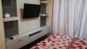 A bedroom with a TV mounted on the wall, a bed with a floral bedspread, and a window with curtains. - Hotel O Apartment Grand Asia Afrika By Prisma (Bandung)
