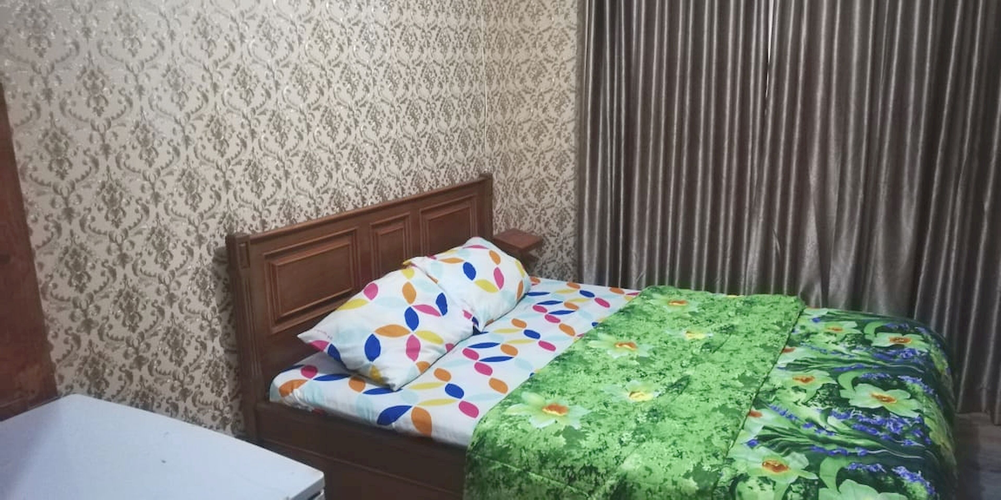 A bedroom with a patterned wallpaper, a wooden headboard, a bed with colorful pillows and a green floral bedspread.